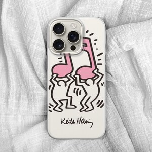 May include: A white phone case featuring a pink musical note design held up by two black and white figures. The case has a black signature at the bottom that reads "Keith Haring". The phone case has a camera module with three lenses.