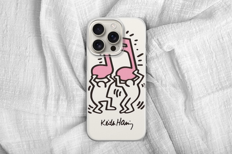 May include: A white phone case featuring a pink musical note design held up by two black and white figures. The case has a black signature at the bottom that reads "Keith Haring". The phone case has a camera module with three lenses.