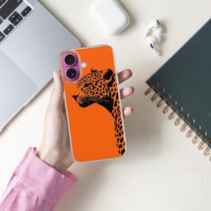 May include: An orange phone case featuring a black leopard design. The case is held near a laptop, a notebook with gold spiral binding, and wireless earbuds. The leopard graphic is a side profile with black spots.