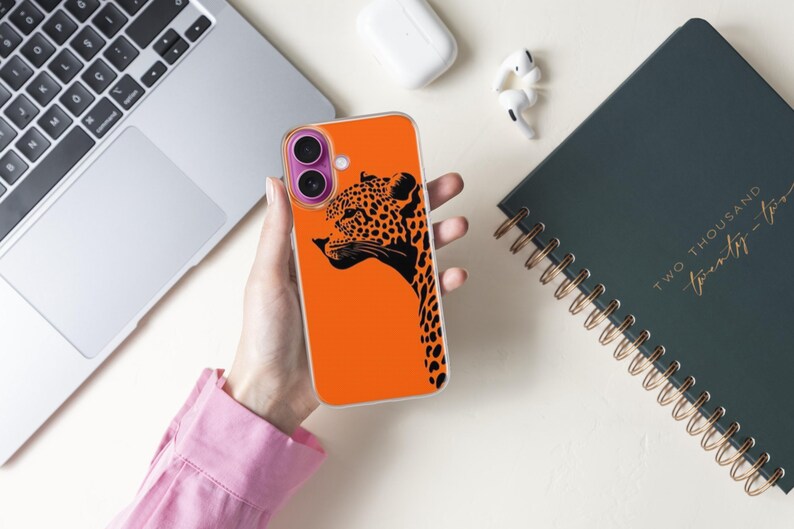 May include: An orange phone case featuring a black leopard design. The case is held near a laptop, a notebook with gold spiral binding, and wireless earbuds. The leopard graphic is a side profile with black spots.