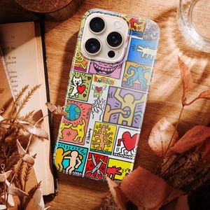 May include: A colorful phone case featuring a pop art design with various figures and shapes in a grid pattern. The case includes a cat with a heart, a globe, and other iconic imagery. The artist's signature, K. Haring, is visible.