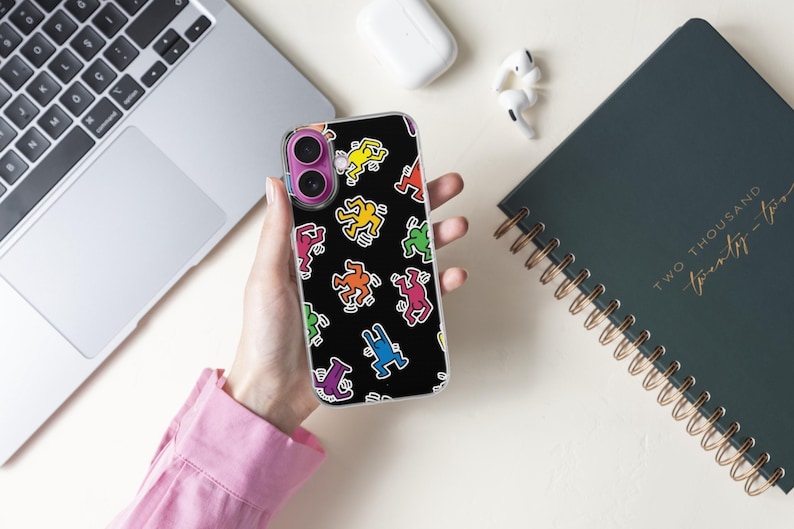 May include: A black phone case featuring a repeating pattern of colorful, stylized figures in various poses. The figures are outlined in black and filled with bright colors like yellow, red, and blue. The case is held in a person's hand.