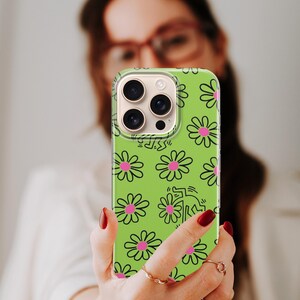 May include: A green phone case with a daisy pattern and a Keith Haring-style figure. The case has a clear border and a camera cutout. The daisies are pink with black outlines. The phone case is being held up.