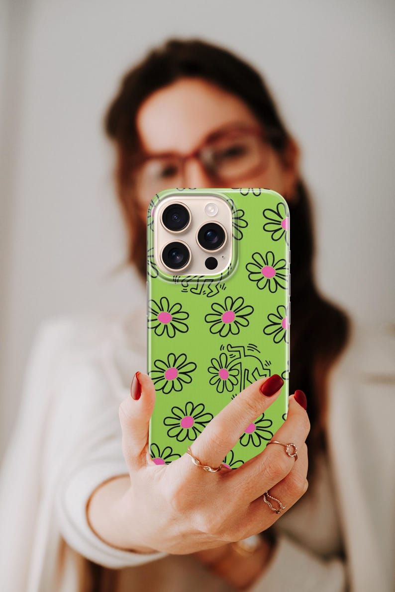 May include: A green phone case with a daisy pattern and a Keith Haring-style figure. The case has a clear border and a camera cutout. The daisies are pink with black outlines. The phone case is being held up.