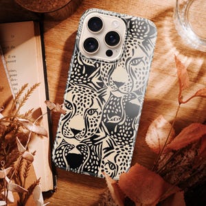 May include: A smartphone case featuring a black and cream-colored leopard face design. The case is on a wooden surface with a book, dried leaves, and a glass of water. The case has a glossy finish and a camera cutout.