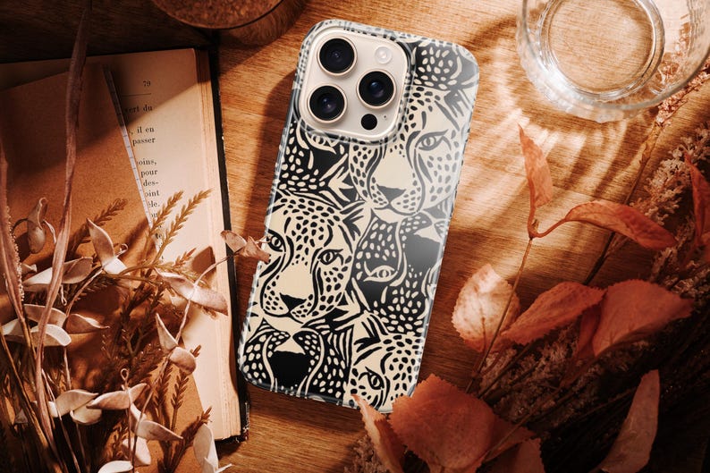 May include: A smartphone case featuring a black and cream-colored leopard face design. The case is on a wooden surface with a book, dried leaves, and a glass of water. The case has a glossy finish and a camera cutout.