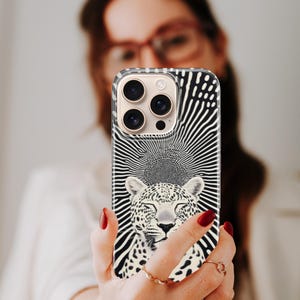 May include: A black and white phone case featuring a leopard design with a sunburst background. The case has a clear border and a camera cutout. The leopard has closed eyes and a serene expression. The case is held up in front of a person.