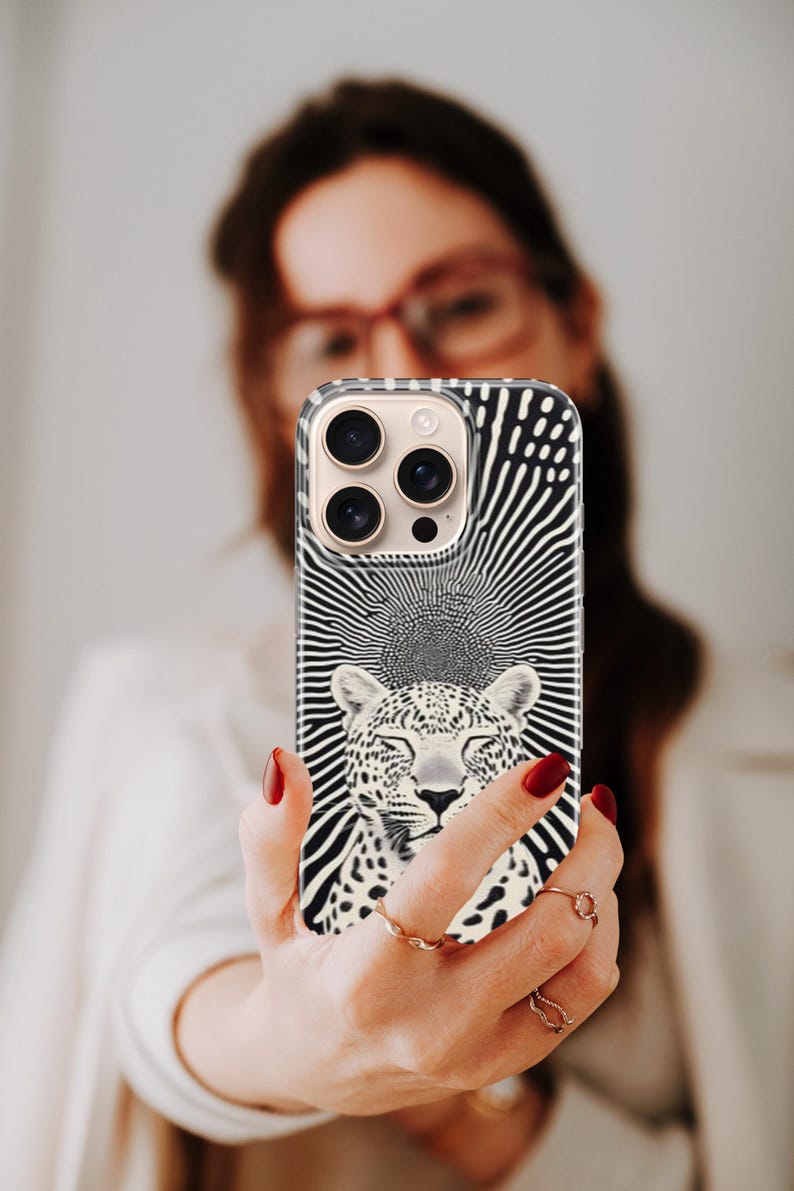 May include: A black and white phone case featuring a leopard design with a sunburst background. The case has a clear border and a camera cutout. The leopard has closed eyes and a serene expression. The case is held up in front of a person.