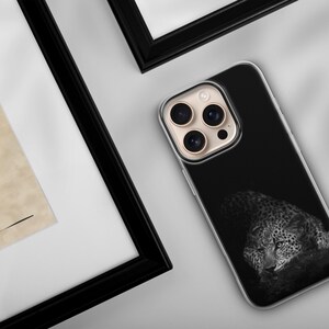 May include: A black phone case featuring a grayscale image of a leopard resting. The phone case is next to a framed artwork with a profile of a face and a red vase. The frame is black.