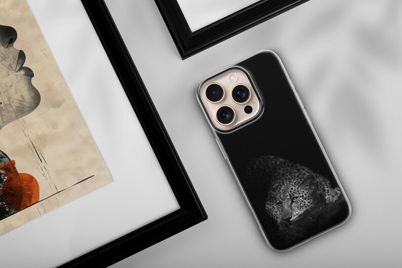 May include: A black phone case featuring a grayscale image of a leopard resting. The phone case is next to a framed artwork with a profile of a face and a red vase. The frame is black.