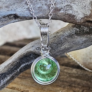May include: A circular pendant featuring a translucent lime green glass cabochon encased in silver-toned wire wrapping. The pendant hangs from a delicate silver-toned metal chain, with the wire forming a decorative coil at the top bail.