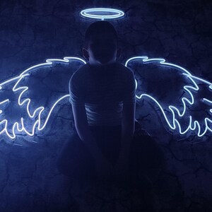May include: A person is silhouetted against a dark blue background, with neon angel wings and a halo. The wings are outlined in bright white neon, and the halo is a simple circle above the head. The overall effect is a striking, ethereal image.