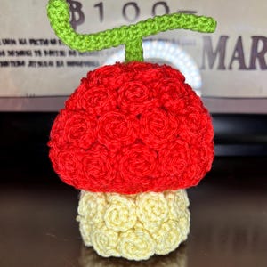 May include: A handmade crochet fruit, featuring a vibrant red top and a light yellow base. The fruit has a green stem. The texture is created by tightly crocheted stitches, giving it a unique, handcrafted appearance.