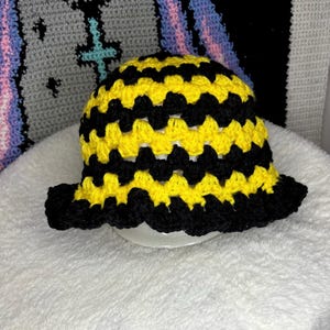 Op de afbeelding: Een handgemaakte gehaakte bucket hat in een opvallend geel en zwart zigzagpatroon. De hoed heeft een geschulpte rand en wordt gepresenteerd op een wit oppervlak. De hoed is een stijlvol accessoire.