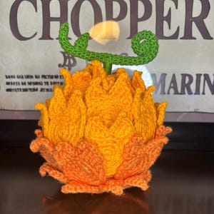 May include: A handmade crochet fruit replica featuring a vibrant orange and yellow textured body with flame-like petal shapes. The top of the fruit has a bright green stem with a distinct curled vine detail, crafted from yarn with visible stitching.