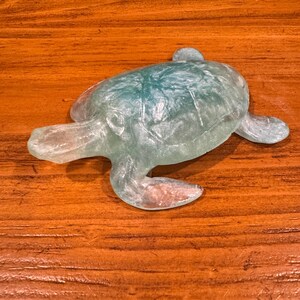 May include: A translucent sea turtle figurine with a blue-green hue. The turtle is made of a clear material, with visible details of its shell, head, and flippers. The figurine is resting on a wooden surface.
