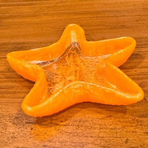 May include: An orange, star-shaped decorative dish. The dish has a glossy finish and a shallow interior. The star dish is sitting on a wooden surface. The dish is a vibrant orange color.