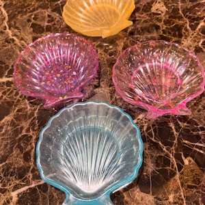 May include: Four shell-shaped dishes made of translucent resin. One dish is yellow, one is light blue, and two are pink. The pink and blue dishes contain embedded glitter particles. Each dish features a scalloped edge and radial ridges.