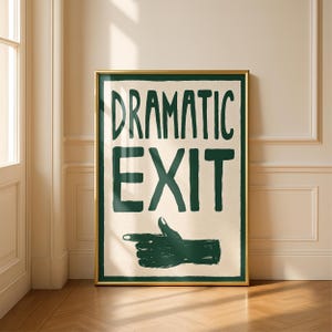 May include: A rectangular wall art print in a gold frame featuring the dark green text DRAMATIC EXIT in a hand-drawn style on a cream background. Below the text is a dark green illustration of a hand pointing to the left with a thumb up.
