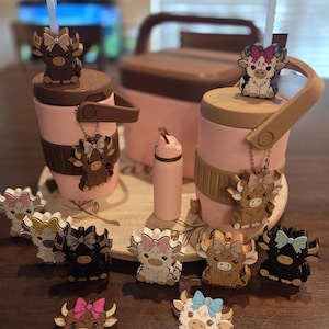 May include: Two light pink insulated tumblers with brown lids and handles, each featuring a straw topper shaped like a highland cow with a bow. Several small highland cow charms with bows in various colors are arranged around the tumblers.
