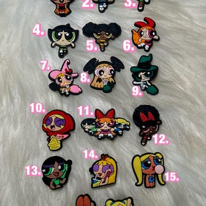 Peut inclure: Une collection de breloques de personnages de dessins animés colorés, présentant divers designs avec des expressions faciales et des tenues distinctes. Les breloques sont disposées sur une surface blanche et moelleuse, mettant en valeur les personnages des Super Nanas.