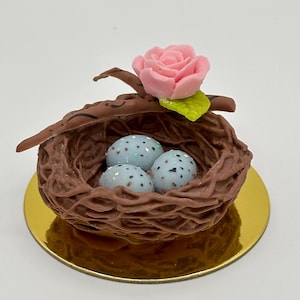 K&ouml;nnte beinhalten: Ein dekoratives Vogelnest mit drei gesprenkelten blauen Eiern, einer rosa Rose und einem gr&uuml;nen Blatt. Das braune Nest steht auf einer goldfarbenen Basis. Das Design ist skurril und detailliert.