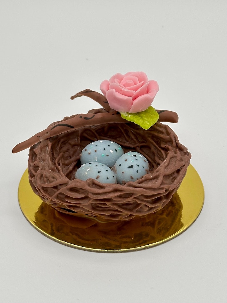 K&ouml;nnte beinhalten: Ein dekoratives Vogelnest mit drei gesprenkelten blauen Eiern, einer rosa Rose und einem gr&uuml;nen Blatt. Das braune Nest steht auf einer goldfarbenen Basis. Das Design ist skurril und detailliert.