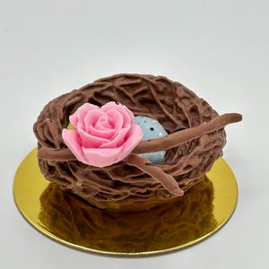 K&ouml;nnte beinhalten: Ein dekoratives Schokoladennest mit einer rosa Rose und einem blau gesprenkelten Ei. Das Nest ist braun und steht auf einer goldfarbenen runden Basis. Die Rose ist ein leuchtendes Rosa und setzt Farbakzente.