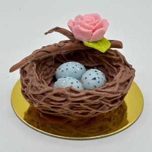 K&ouml;nnte beinhalten: Ein dekoratives Vogelnest mit drei gesprenkelten blauen Eiern. Das braune Nest ist mit einer rosa Rose und einem gr&uuml;nen Blatt gekr&ouml;nt. Das Nest steht auf einer goldfarbenen runden Basis und schafft ein charmantes, von der Natur inspiriertes Design.