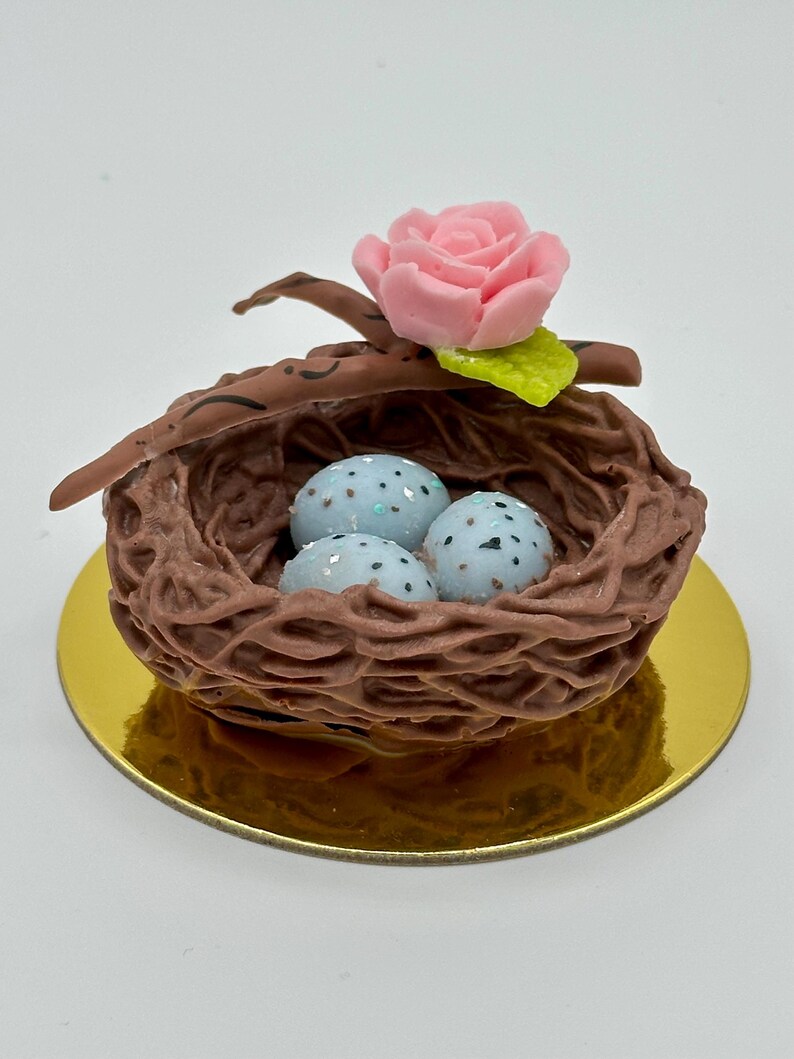 K&ouml;nnte beinhalten: Ein dekoratives Vogelnest mit drei gesprenkelten blauen Eiern. Das braune Nest ist mit einer rosa Rose und einem gr&uuml;nen Blatt gekr&ouml;nt. Das Nest steht auf einer goldfarbenen runden Basis und schafft ein charmantes, von der Natur inspiriertes Design.