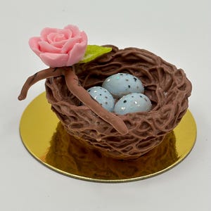 K&ouml;nnte beinhalten: Ein dekoratives Vogelnest mit drei gesprenkelten blauen Eiern, einer rosa Rose und einem gr&uuml;nen Blatt. Das Nest ist braun und steht auf einer goldfarbenen runden Basis. Ein bezaubernder, handgefertigter Artikel.