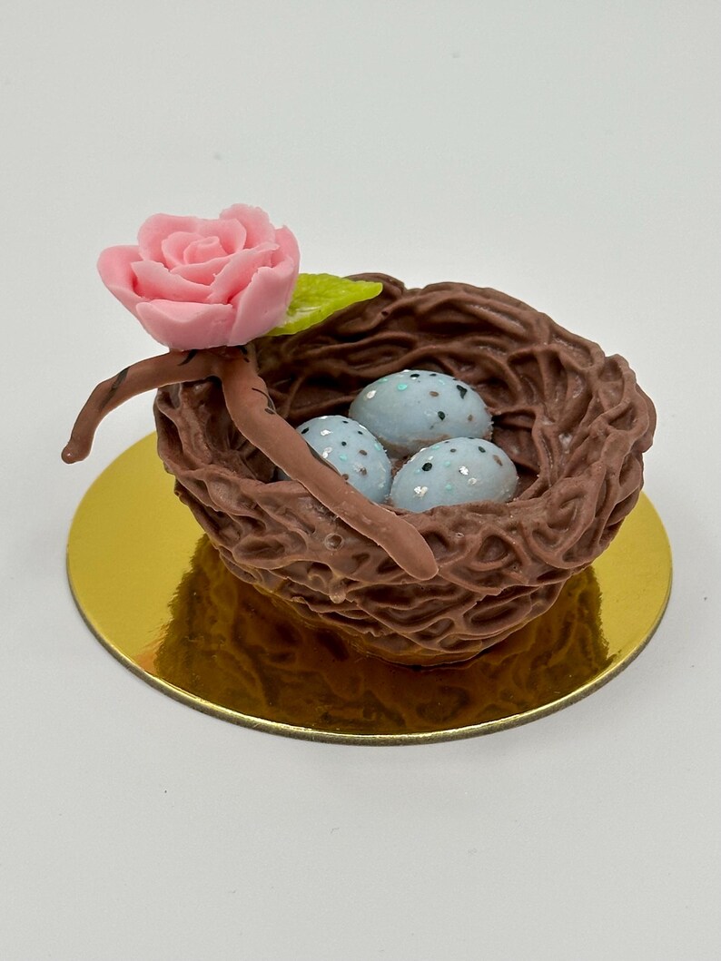 K&ouml;nnte beinhalten: Ein dekoratives Vogelnest mit drei gesprenkelten blauen Eiern, einer rosa Rose und einem gr&uuml;nen Blatt. Das Nest ist braun und steht auf einer goldfarbenen runden Basis. Ein bezaubernder, handgefertigter Artikel.