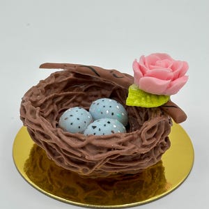K&ouml;nnte beinhalten: Ein dekoratives Vogelnest mit drei gesprenkelten blauen Eiern, einer rosa Rose und einem gr&uuml;nen Blatt. Das Nest ist braun und steht auf einer goldfarbenen runden Basis. Ein charmantes, naturbezogenes Deko-Objekt.