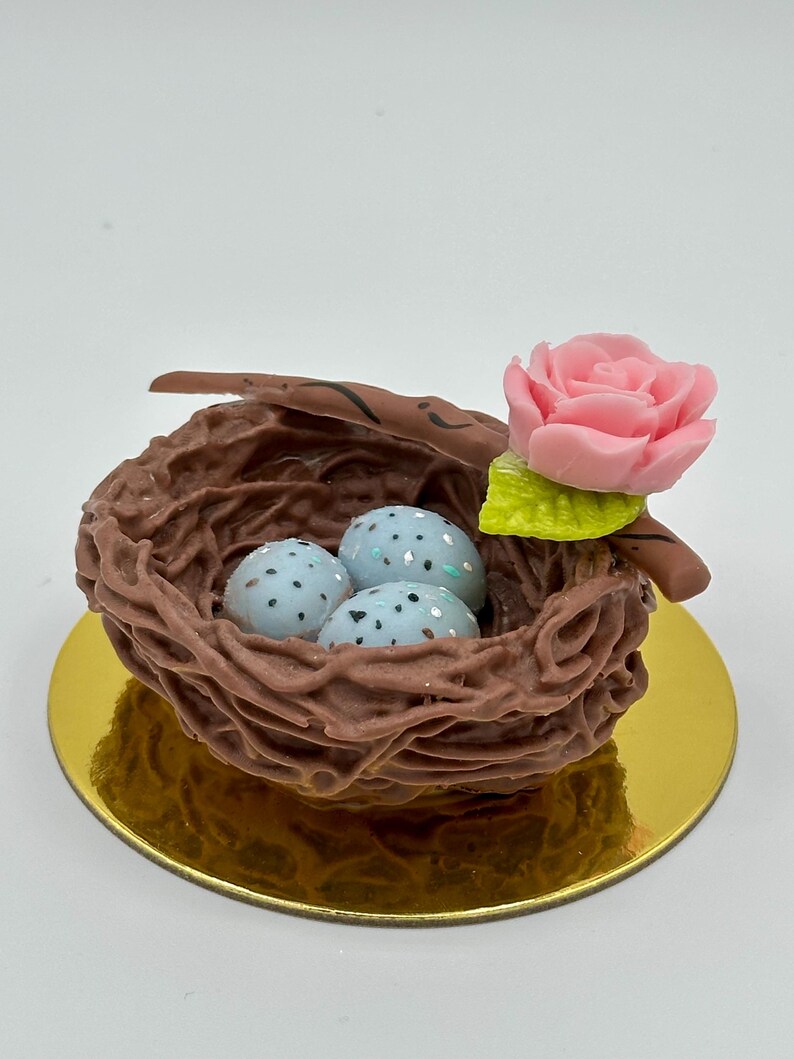 K&ouml;nnte beinhalten: Ein dekoratives Vogelnest mit drei gesprenkelten blauen Eiern, einer rosa Rose und einem gr&uuml;nen Blatt. Das Nest ist braun und steht auf einer goldfarbenen runden Basis. Ein charmantes, naturbezogenes Deko-Objekt.