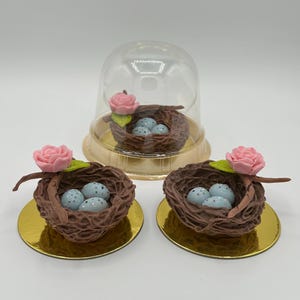 K&ouml;nnte beinhalten: Drei dekorative Vogelnester mit blau gesprenkelten Eiern. Jedes Nest ist braun und hat eine rosa Rose und einen gr&uuml;nen Blattakzent. Ein Nest befindet sich unter einer transparenten Kunststoffkuppel, alle auf goldenen kreisf&ouml;rmigen Basen.