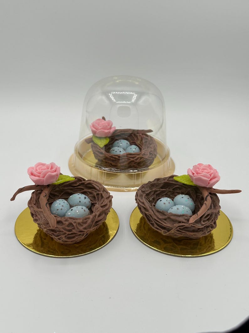 K&ouml;nnte beinhalten: Drei dekorative Vogelnester mit blau gesprenkelten Eiern. Jedes Nest ist braun und hat eine rosa Rose und einen gr&uuml;nen Blattakzent. Ein Nest befindet sich unter einer transparenten Kunststoffkuppel, alle auf goldenen kreisf&ouml;rmigen Basen.