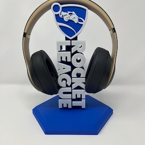 Puede incluir: Auriculares sobre un soporte azul y blanco con la inscripción "ROCKET LEAGUE" en blanco. El soporte tiene una base azul y un logotipo blanco con un coche. Los auriculares son negros con una diadema dorada.