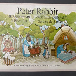 May include: An illustration of a Peter Rabbit nurseryware 3-piece set. The image depicts cartoon rabbits, trees, and a cottage. Text includes "Peter Rabbit," "Nurseryware," and "3 Piece Set." The set includes a cereal bowl, mug, and plate.