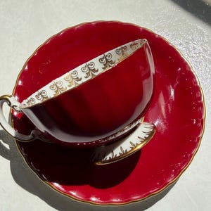 May include: A deep red teacup and matching saucer set featuring a scalloped edge and a gold-coloured rim. The interior of the teacup is white with a decorative gold filigree pattern along the upper border, and the handle is white with gold trim.