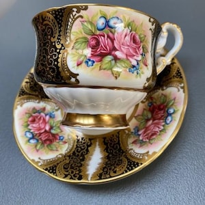 May include: A vintage teacup and saucer set. The cup and saucer feature pink roses, blue flowers, and gold detailing. The cup has a black and gold patterned design. The set is displayed on a light blue surface.