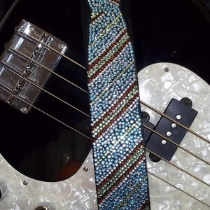 Puede incluir: Un bajo negro con una golpeador blanco y una correa de guitarra de lazo de cristal azul y verde con la palabra "ERNIE" en la correa.