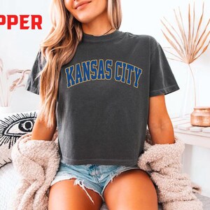 May include: Dark gray t-shirt with the text "KANSAS CITY" in blue and gold lettering. The shirt is a crew neck and appears to be made of a soft material. The word "PEPPER" is in red in the upper left corner.