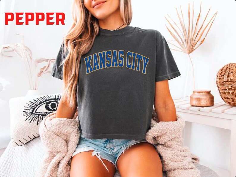 May include: Dark gray t-shirt with the text "KANSAS CITY" in blue and gold lettering. The shirt is a crew neck and appears to be made of a soft material. The word "PEPPER" is in red in the upper left corner.