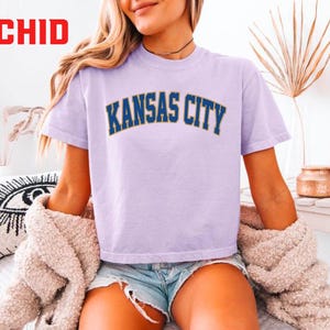 May include: A light purple t-shirt with "KANSAS CITY" printed in navy blue and gold lettering. The word "ORCHID" is in red in the upper left corner. The shirt is paired with denim shorts and a beige sweater.