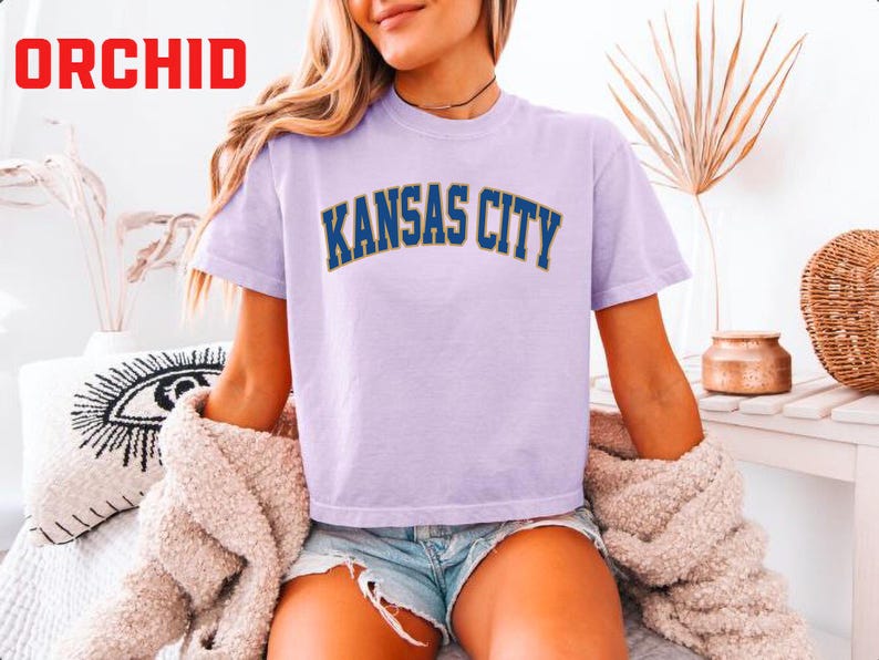 May include: A light purple t-shirt with "KANSAS CITY" printed in navy blue and gold lettering. The word "ORCHID" is in red in the upper left corner. The shirt is paired with denim shorts and a beige sweater.