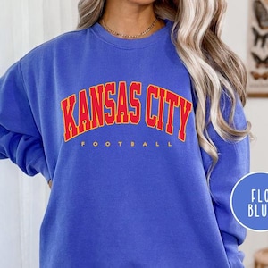 May include: A blue sweatshirt with the text "KANSAS CITY FOOTBALL" in red and yellow lettering. The sweatshirt is a solid blue color and has long sleeves. A blue circle with the text "FLO BLUE" is in the lower right corner.
