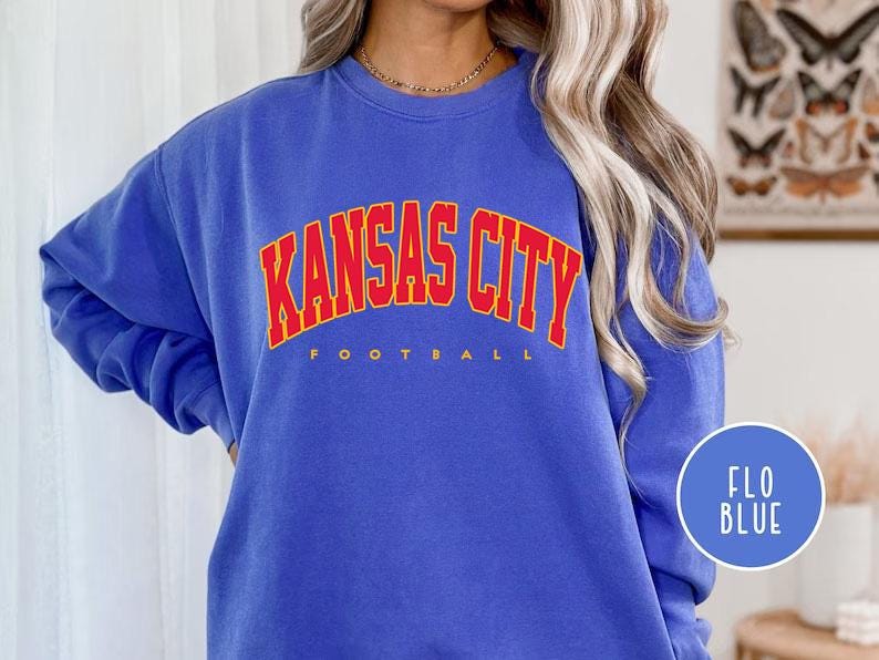 May include: A blue sweatshirt with the text "KANSAS CITY FOOTBALL" in red and yellow lettering. The sweatshirt is a solid blue color and has long sleeves. A blue circle with the text "FLO BLUE" is in the lower right corner.