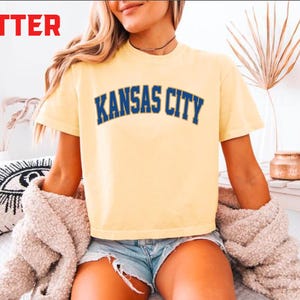 May include: A light yellow t-shirt with the text "KANSAS CITY" in blue lettering. The shirt is worn with denim shorts and a beige sweater. The word "BUTTER" is in red in the top left corner.