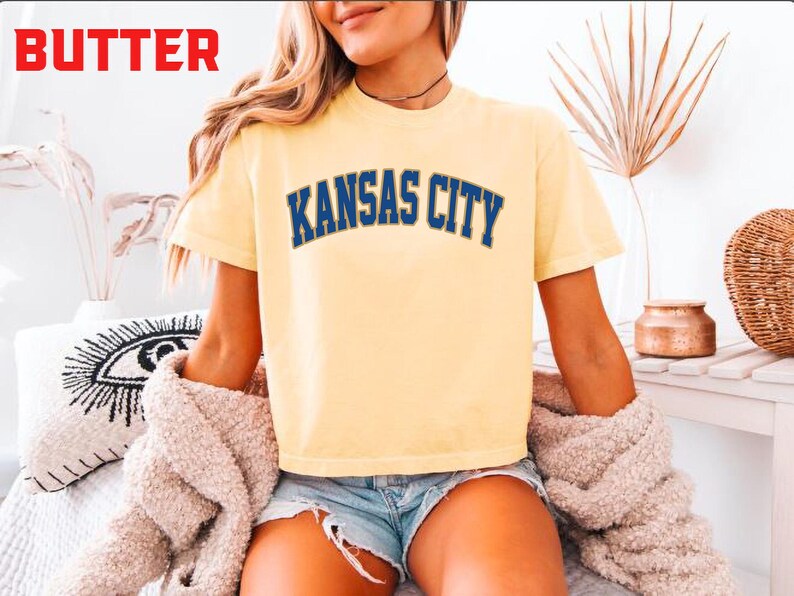 May include: A light yellow t-shirt with the text "KANSAS CITY" in blue lettering. The shirt is worn with denim shorts and a beige sweater. The word "BUTTER" is in red in the top left corner.