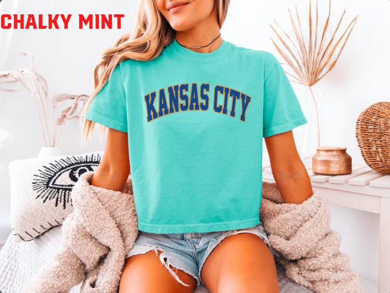 May include: A turquoise t-shirt with the text "KANSAS CITY" in navy blue arched lettering. The words "CHALKY MINT" are in red at the top left. The shirt is short-sleeved and worn with denim shorts.